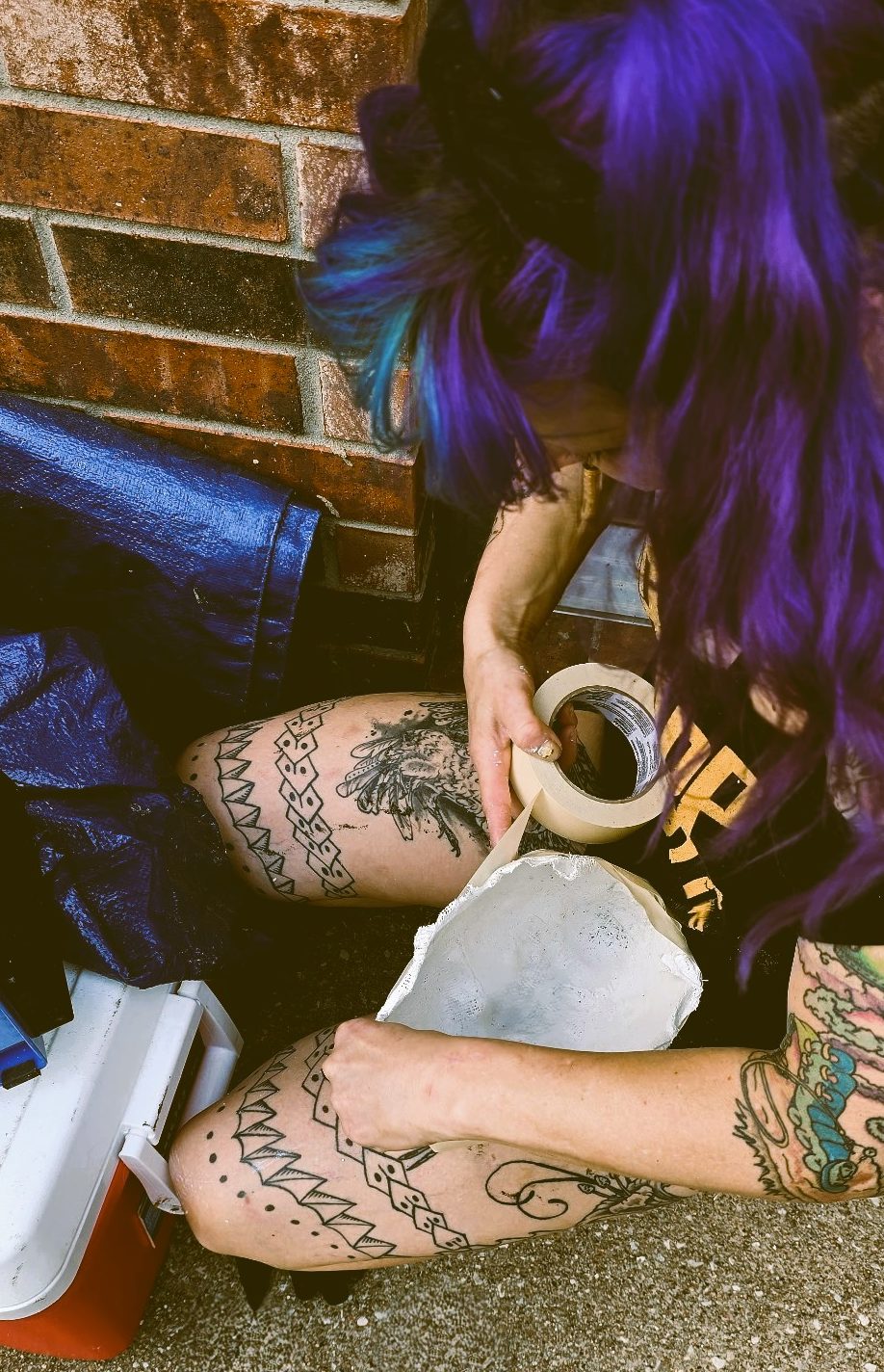 Artist sculpting a mask by hand, seated with focus and intention, against a brick wall