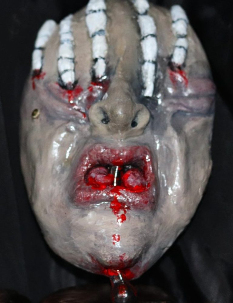 Front view of “SPLIT” mask sculpture with split tongue, bike brake lever, and skeletal hand detail