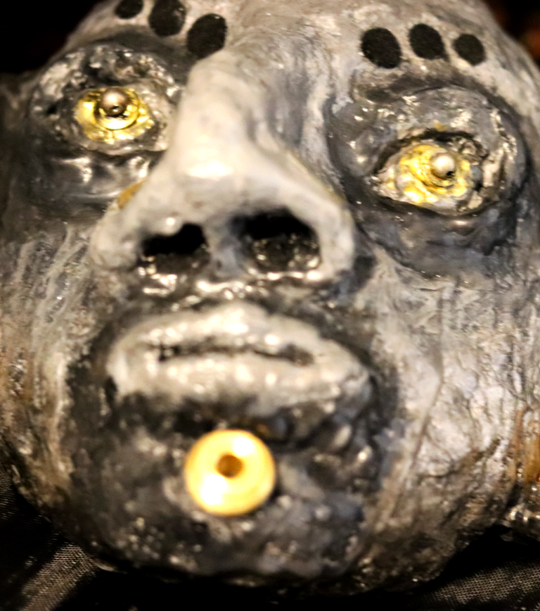 Mr. Money – Frontal View Frontal view of the Mr. Money sculpture showing yellow-gold eyes, tribal facial features, and gold piercings